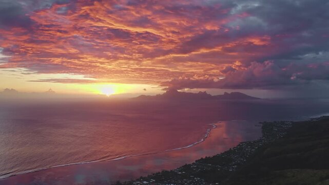 Moorea coastline at dusk, vivid pink, orange and purple sky reflected on calm Pacific waters with silhouetted mountains and tranquil tropical beach. Natural landscape. Aerial view drone flight