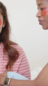 Vertical video: Approaching woman joining friend doing skincare on bed, holding pillow with patches