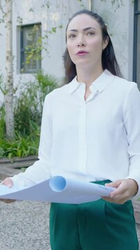 Vertical video: Pulling back camera, woman white blouse checking plans for site in courtyard, phone