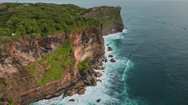 Steep limestone cliffs of Bali's Bukit Peninsula plunge into turquoise Indian Ocean waters green vegetation, waves crash on rugged rocky shores under a bright sky. Aerial view drone flight panorama