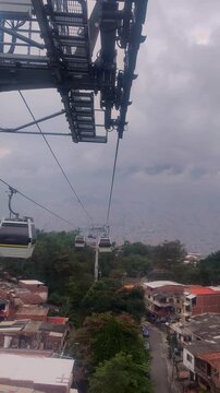 Metrocable cable car tower overlooking a dense hillside neighborhood in Medellin, Colombia, with trees, rooftops, and a cloudy mountain backdrop viewed from inside a gondola cabin.