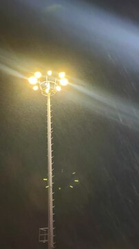 Tall stadium floodlight glows in heavy rain and mist at night, creating dramatic beams and lens flare. Dark atmospheric scene with intense illuminated droplets and urban outdoor mood.