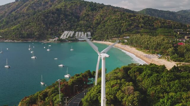 Wind turbine spinning its blades, generating clean sustainable energy above a tropical bay with yachts and Nai Harn beach, surrounded by green hills in Phuket, Thailand. Aerial view drone shot