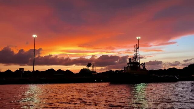 Vibrant orange and purple sunset sky over calm water, with dark silhouetted shoreline, tall light pole, and reflections creating a dramatic evening seascape.