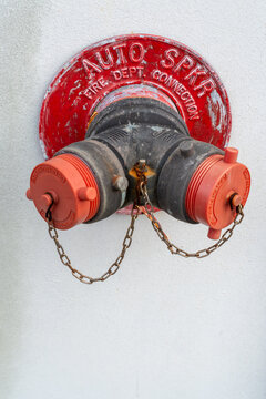 Vertical photo of a fire department connection mounted on a white wall, featuring two weathered metal pipe outlets with red caps secured by chains and red safety signage above