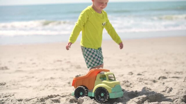 Young boy in bright green shirt and patterned shorts bends over a toy truck on sandy beach, filling it with sand as ocean waves lap at the shore in the background