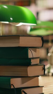 Vertical format shot: Close up of stacked books on desk in quiet university library with warm lighting and green lamp, educational environment with bookshelves and study materials.