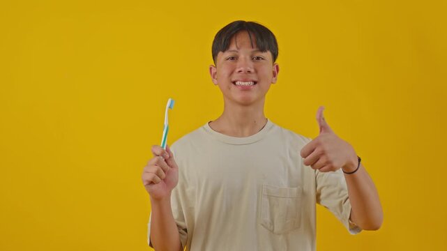 Happy Asian teenager holding toothbrush and showing thumb up against yellow background