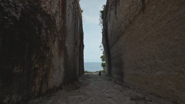 Man on scooter riding narrow dirt path trail carved between tall cliffs, traveling towards expansive blue ocean and a sandy beach under a bright sky, symbolizing adventure and tropical escape, Bali