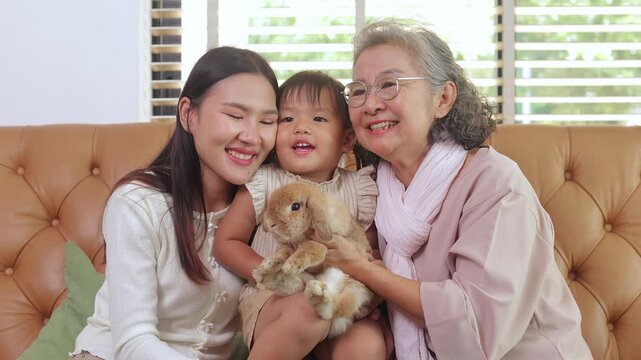 Happy multi-generational Asian family with grandmother, mother and child smiling while hugging a cute pet rabbit together on a brown sofa in their cozy living room. pet concept