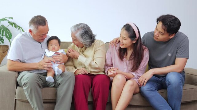 Happy multi-generational Asian family sitting on a sofa. Smiling grandparents, parents, and a cute baby enjoying quality time together at home, sharing love and warm memories.