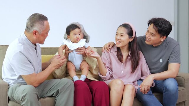 Happy multi-generational Asian family sitting on a sofa. Smiling grandparents, parents, and a cute baby enjoying quality time together at home, sharing love and warm memories.
