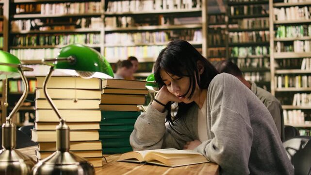 Tired woman student leans on her hand while reading open book under green lamp, surrounded by tall stacks of books in a warm academic library at wooden desk, Focus on self study, with study session.