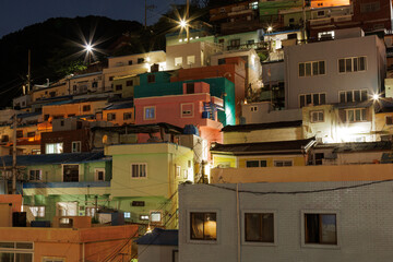 Fototapeta premium Brightly illuminated colorful houses on a hillside at night in Busan, South Korea. Dense residential neighborhood with glowing windows, city lights and mountain backdrop.