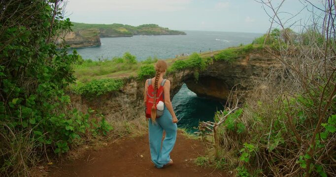 Woman backpacker descending dirt path towards Broken Beach natural arch view Nusa Penida Bali