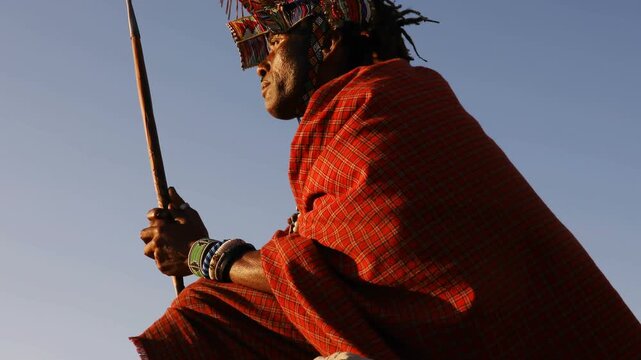 Maasai Warrior in Traditional Clothing Sitting on a Rock with a Spear Against Blue Sky Background