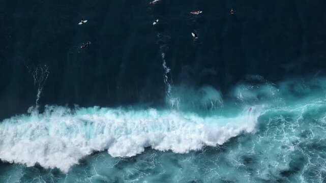 Surfers are riding waves and waiting in the crystal blue ocean water, enjoying the popular water sport activity on the scenic Bukit Peninsula in Uluwatu, Bali. Aerial view drone flight