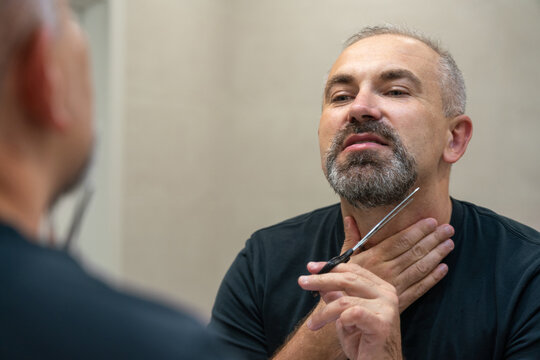 Middle-aged handsome man using scissors to cut his beard a litlie and fix the shape