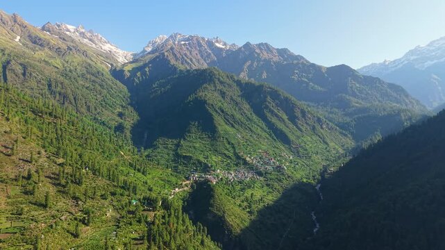 Grahan Village nestled among dense pine forests, steep green slopes, and snow capped Himalayan mountains near Kasol in Parvati Valley, Himachal Pradesh, India. Aerial panoramic drone dolly in