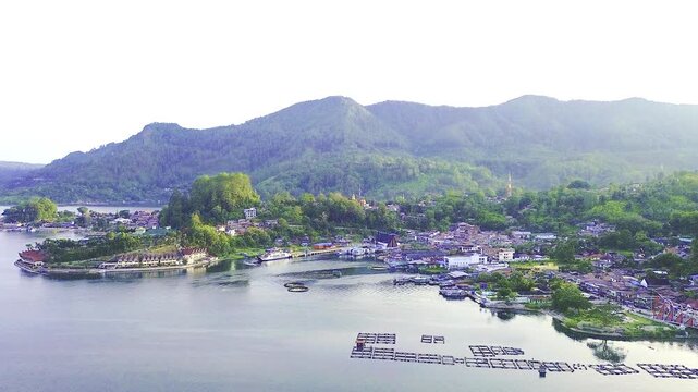 Scenic drone aerial view of a quiet lakeside village at Lake Toba, Indonesia, featuring fish farming cages on the water, lush hills in the background, and a serene natural environment