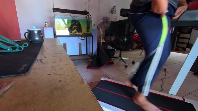 Video of Person walks barefoot on treadmill at home. Home gym setup includes table, fish tank, and mat. No visible emotion on person's face. Image captures casual workout at home. Good for fitness, li