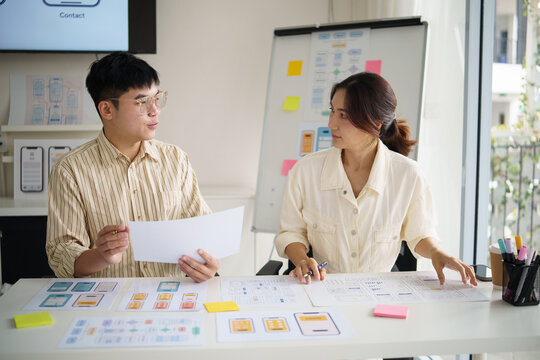 Two UX designers collaborating on mobile app wireframes, reviewing paper prototypes and user interface layouts in a bright office.