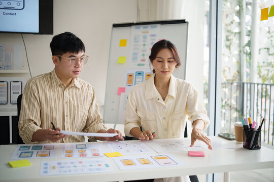 Two UX designers collaborating on mobile app wireframes, reviewing paper prototypes and user interface layouts in a bright office.