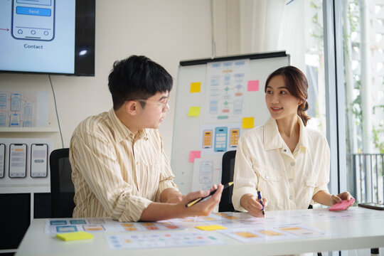Two UX designers collaborating on mobile app wireframes, reviewing paper prototypes and user interface layouts in a bright office.