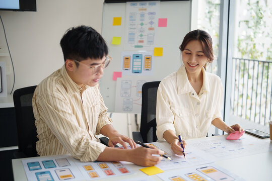 Two UX designers collaborating on mobile app wireframes, reviewing paper prototypes and user interface layouts in a bright office.