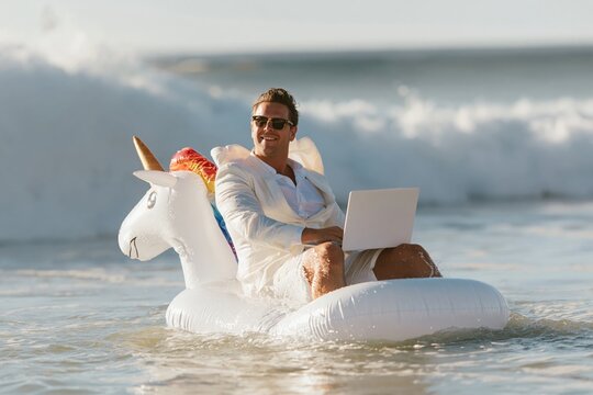A man in a white suit works on a laptop while floating on an inflatable unicorn in the ocean, for remote work humor and digital nomad lifestyle content