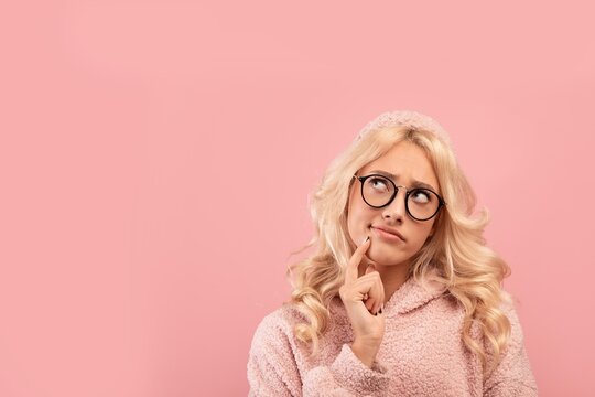 Contemplative Young Woman In Pink Sweater With Glasses Thinking, Holding Finger On Chin While Pondering Ideas Against Solid Pink Background. Concept Of Creativity And Inspiration.