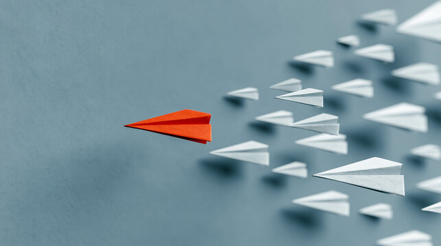 Red Paper Plane Leading a Flock: An orange paper airplane soars ahead of a cluster of white paper airplanes against a muted blue background, symbolizing leadership and innovation.