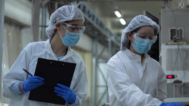 Quality control team using digital tablet and clipboard for production line audit in modern plant, Female scientists wearing face masks and safety goggles working in high-standard processing facility