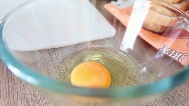 Cooking process showing three raw eggs being opened into a transparent bowl on a wooden table.

