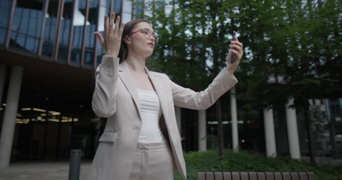Young Caucasian businesswoman or student or intern in a business suit and glasses in an urban setting among trees and modern buildings making a video call on the phone