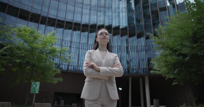 A young Caucasian businesswoman or student or intern in a business suit and glasses in an urban setting among trees and modern buildings stands with her arms crossed - orbital shot