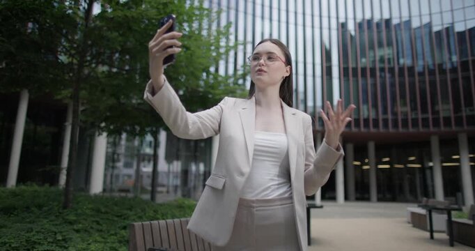 Young Caucasian businesswoman or student or intern in a business suit and glasses in an urban setting among trees and modern buildings making a video call on the phone