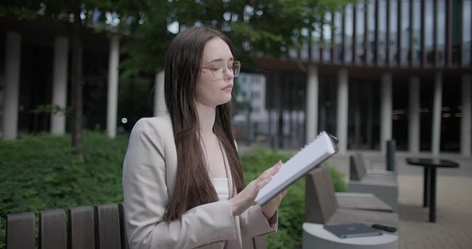 A young Caucasian businesswoman or student or intern in a business suit and glasses in an urban setting among trees and modern buildings sits on a bench and flips through a notebook or reads a book