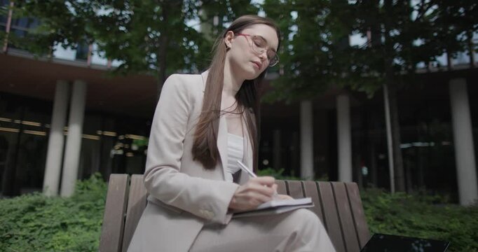 A young Caucasian businesswoman or student or intern in a business suit and glasses in an urban setting among trees and modern buildings sits on a bench and works on a laptop making notes in notebook