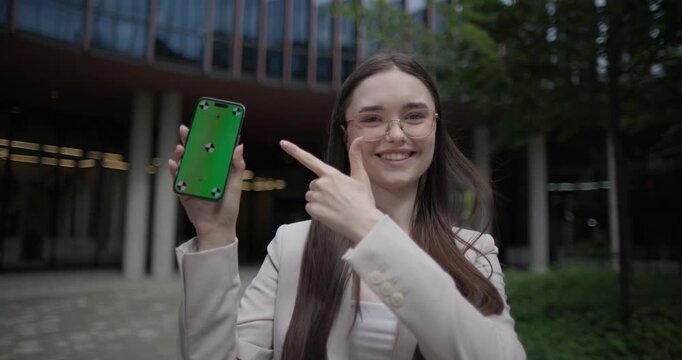 A young Caucasian businesswoman or student or intern in a business suit and glasses in an urban setting among trees and modern buildings presents and shows something on a phone screen and gestures
