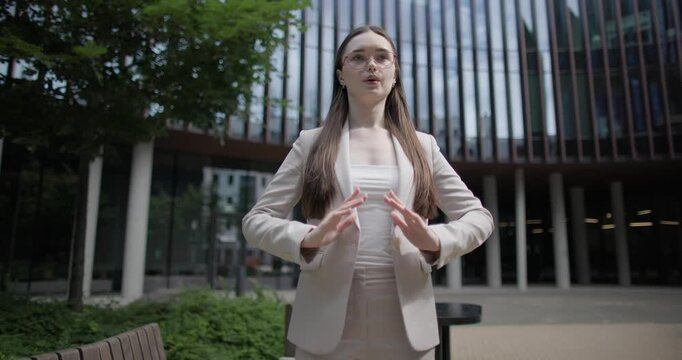 A young Caucasian businesswoman or student or intern in a business suit and glasses in an urban setting among trees and modern buildings does breathing exercises and meditates to relieve stress 