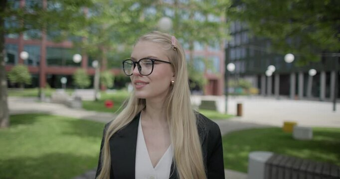 A young Caucasian woman who studies daily wearing glasses stands in an urban setting among houses and trees and looks at the camera.