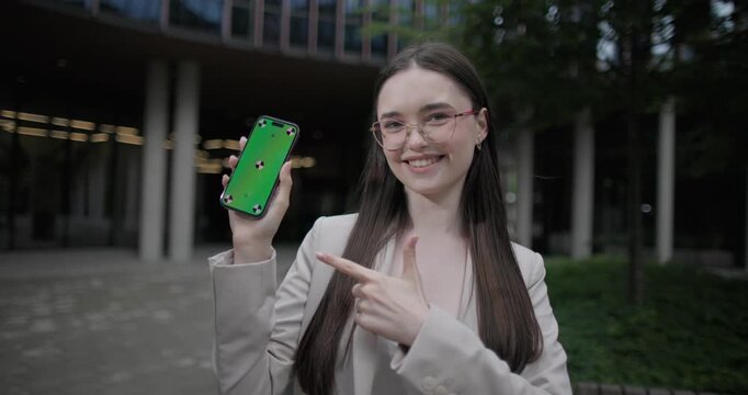 A young Caucasian businesswoman or student or intern in a business suit and glasses in an urban setting among trees and modern buildings presents and shows something on a phone screen and gestures