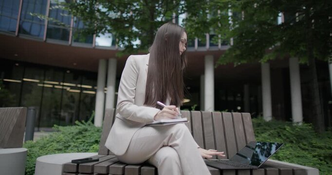 A young Caucasian businesswoman or student or intern in a business suit and glasses in an urban setting among trees and modern buildings sits on a bench and works on a laptop making notes in notebook