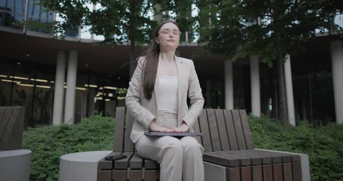 A young Caucasian businesswoman or student or intern in a business suit and glasses in an urban setting among trees and modern buildings sits on a bench and closes the lid of a laptop 