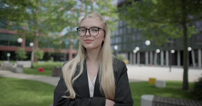 A young Caucasian woman who studies daily wearing glasses stands in an urban setting among houses and trees, crosses her arms over her chest and looks at the camera.