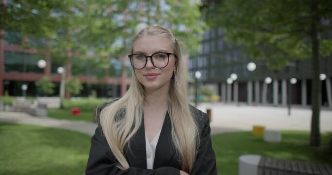 A young Caucasian woman who studies daily wearing glasses stands in an urban setting among houses and trees, crosses her arms over her chest and looks at the camera.