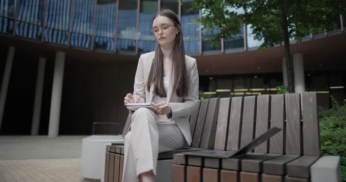 A young Caucasian businesswoman or student or intern in a business suit and glasses in an urban setting among trees and modern buildings sits on a bench and works on a laptop making notes in notebook
