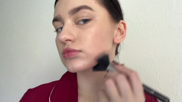 Young woman applying face contouring product with a soft cosmetic brush, emphasizing her prominent cheekbones and sculpting her jawline, demonstrating a personal beauty routine indoors.