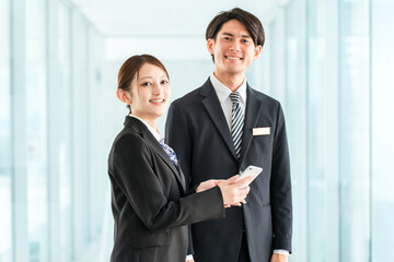 Fototapeta premium A businessman and a woman in suits (hotel staff, consultant) talking while looking at their smartphones in an office hallway.